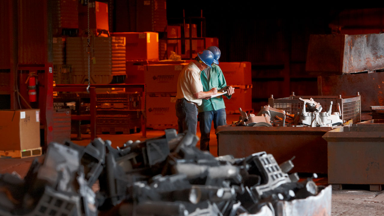 An image of two men in an industrial warehouse, both wear hardhats as they look over paperwork together.